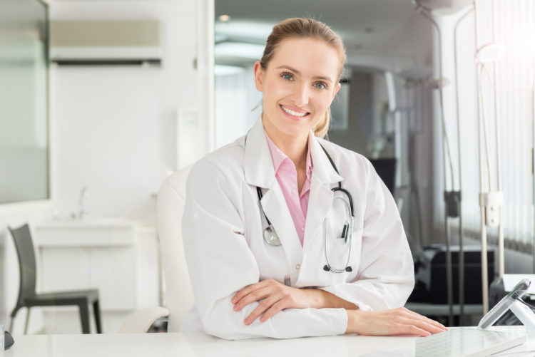 Horizontal color image of pretty female doctor at office sincerely smiling at camera. Wearing white lab coat and stethoscope around neck.