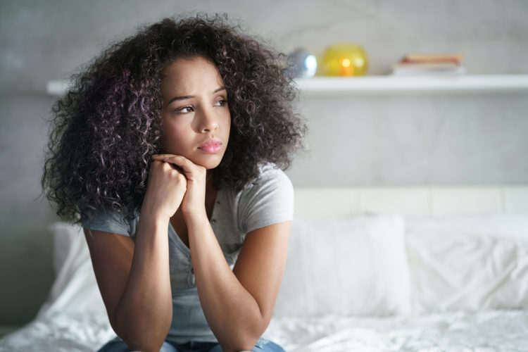 Lonely young latina woman sitting on bed. Depressed hispanic girl at home, looking away with sad expression.