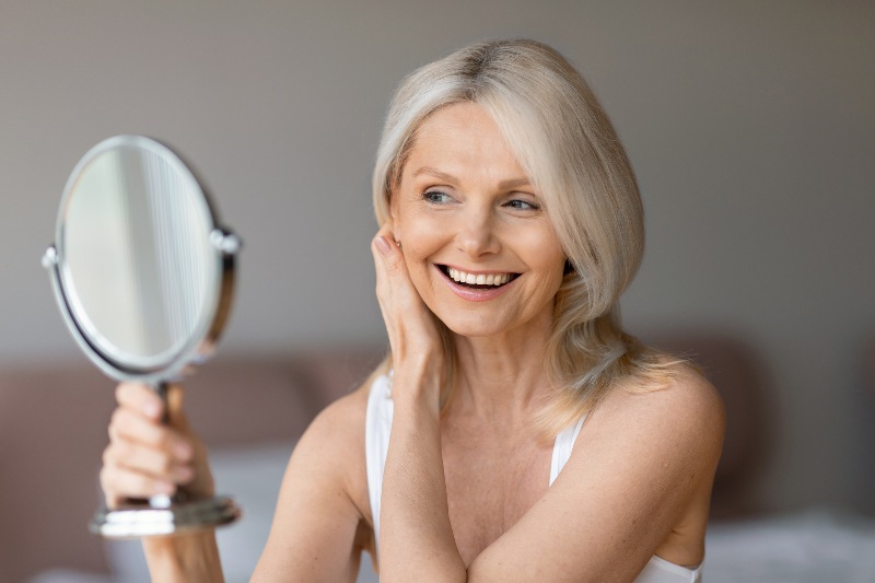 A mature woman looking at her skin in a handheld mirror by Swinyer Woseth Dermatology