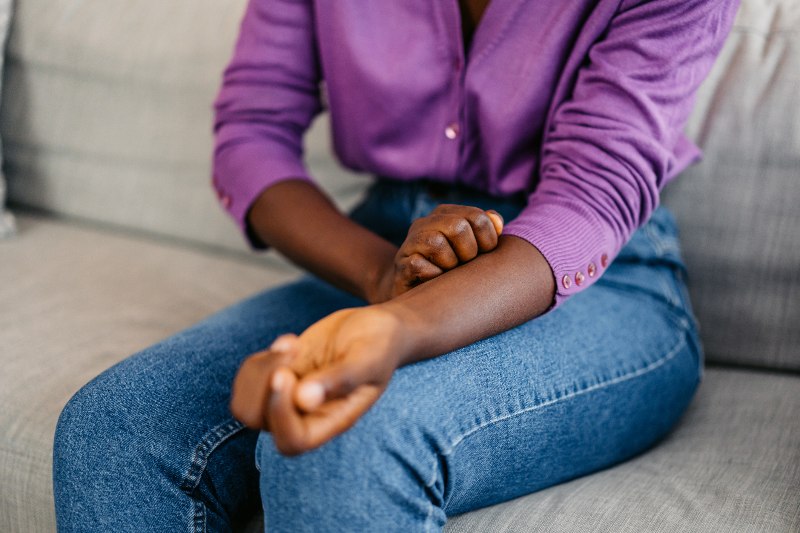 A woman sitting on the couch and scratching her forearm by Swinyer Woseth Dermatology